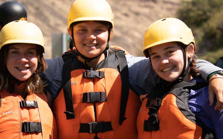 three outward bound students wearing life jackets and helmets wrap their arms around each other's shoulders and pose for the camera 